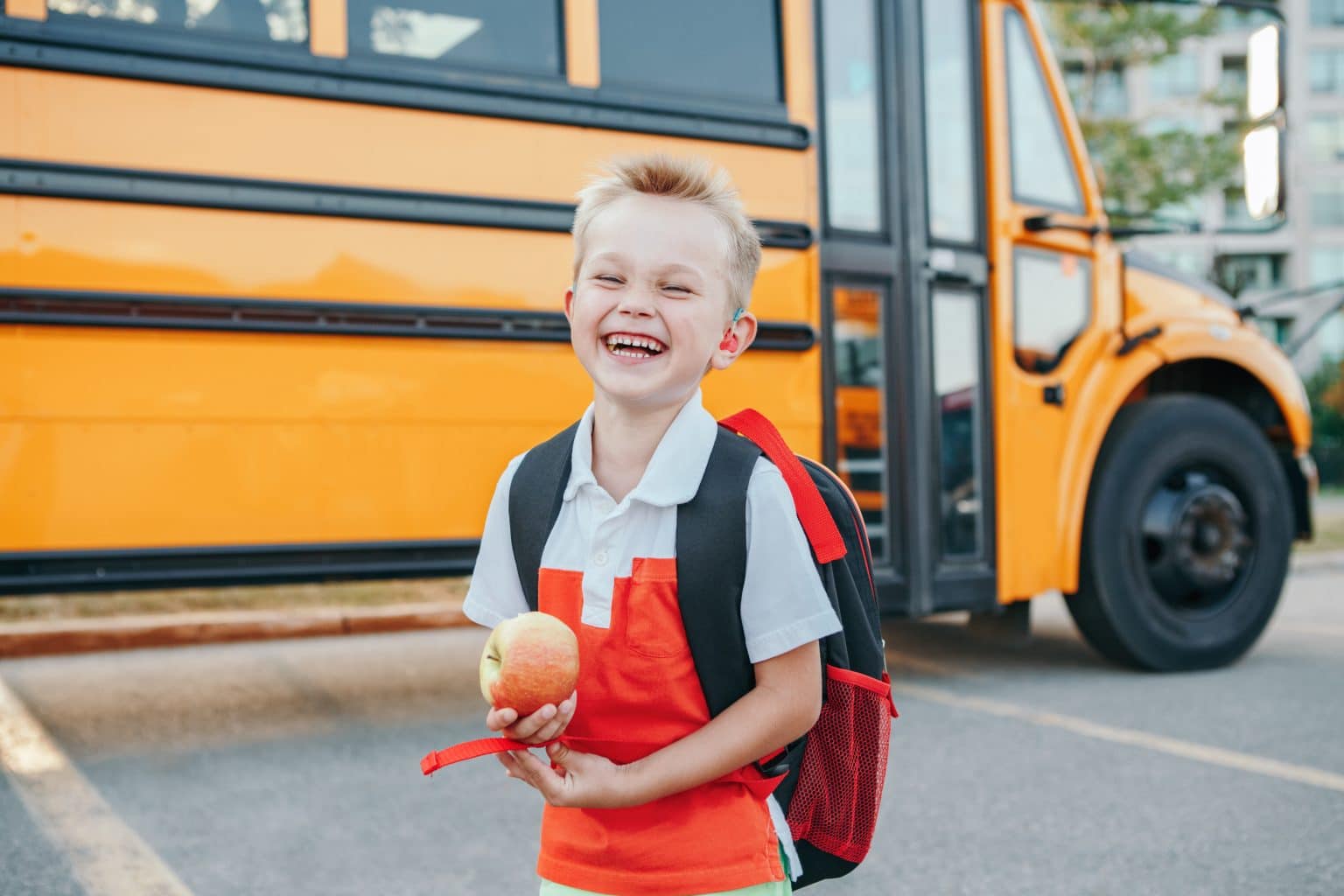 Smiling child wearing a backpack and holding an apple. There is a schoolbus parked behind them. 